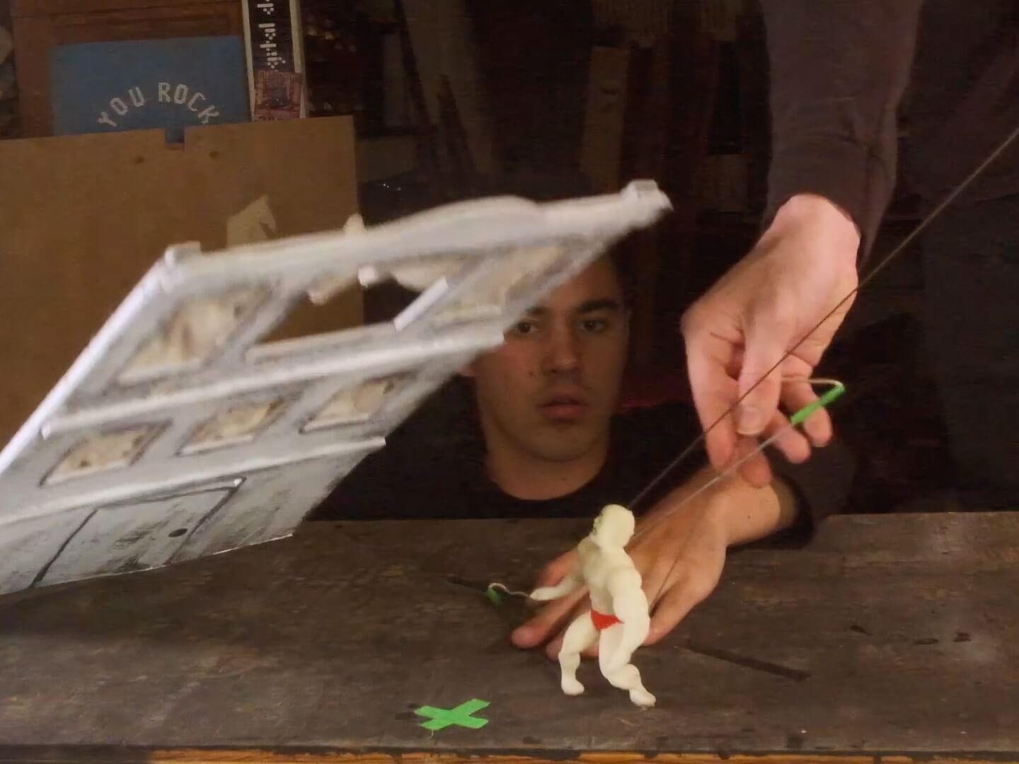 In a spoof of Buster Keaton's famous falling house movie stunt, where the actor stands in exactly the right spot so that the facade of a house falls all around him and leaves him standing unharmed in the space left by an open attic window, a 4-inch-tall yellow foam puppet crouches and looks up at a crudely built white styrofoam facade of a three-story house that's falling and appears about to crush him. A young man dressed in black, his face just above the level of the table, watches the scene and uses his left hand to help manipulate the puppet, while a hand from an unseen second puppeteer reaches intro frame from the top right corner to also manipulate the puppet with a set of metal rods.