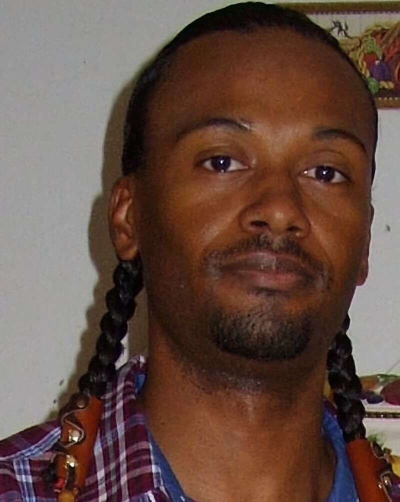 The photo depicts Nant'a (Chief) Cougar Goodbear, an Indigenous American of Canneci Tinne (Apache) and La Kreyol. His head leans slightly to the left, as he looks at the camera with a slight smile. His hair is slicked back into two braids hanging from either side of his head.