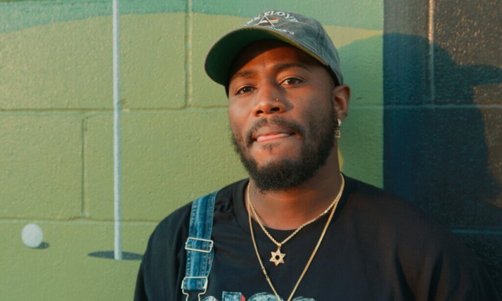 The photo shows Devonta Ravisee, a Black man with brown skin, in front of a historic mural on 4th Avenue in Birmingham. The wall depicts a golf ball approaching a hole on a golf course, and the artist stands in the center looking off-camera with his mouth closed. He is wearing a black shirt, a gray hat, and two gold necklaces.
