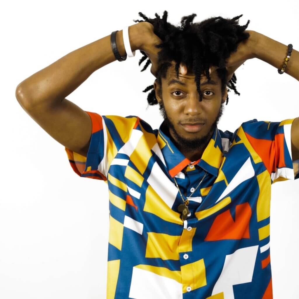 Jahman Hill, a spry youngish Black man with brown skin stares into the camera, holding his wild curly hair up in his hands. He is wearing a swanky multi-colored shirt. Blue, yellow, white, and red in quirky, blocky patterns and a gold necklace with an Africa-shaped pendant. Behind the camera is Janavian Young, one of the greatest documentarians of her generation. She is Black, a woman, and steady of hand. Her camera sports a sunflower strap, both her and Jahman's favorite flower.