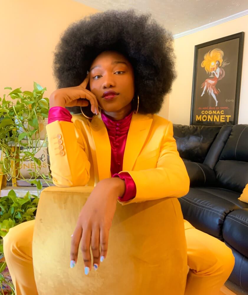 The photo depicts Jazzie Jelks, a young Black woman with medium brown skin and an afro. She poses seated on a velvet yellow chair with its back facing the camera, propping her face on one hand, and draping her arm across the chair and wearing a yellow suit and pink, high-collar blouse. There is a black sofa, a 1920s-themed painting of a woman on the wall, and vining plants in the background.