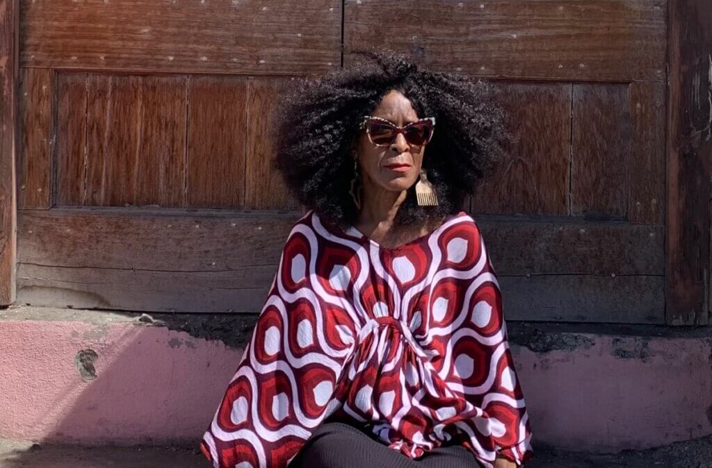 A Black woman with big hair, purple shades, and an afro-pick earring dressed in a maroon, red-orange and periwinkle flowy blouse sits on the stairs in the archway of a pink stone building in front an antique teak wooden door.