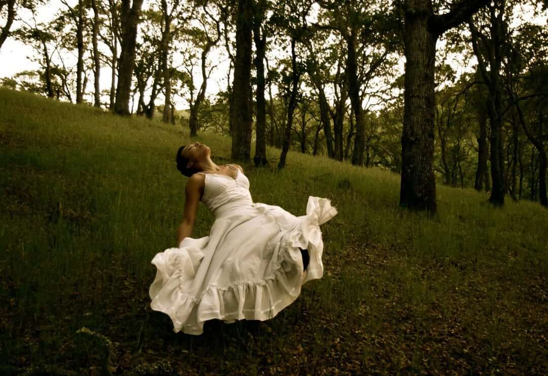 An Afro-Indigenous dancer with dark hair and brown skin floats through a lush, wooded landscape. Immersed in ancestral rites, their body leans backward in a posture of simultaneous surrender to the land and preparation for flight.