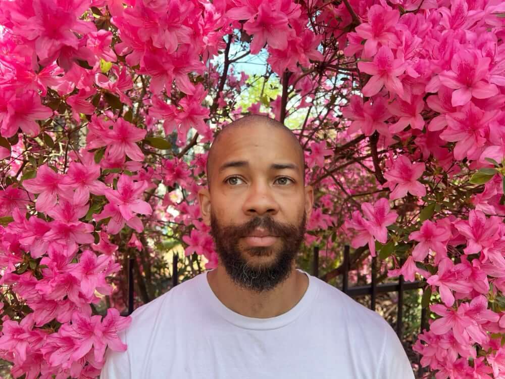 Sean G. Clark The photo shows a medium-skinned African American man in a white T-shirt. He has a shaven head and dark beard and stands immersed in an azalea bush full of pink blossoms. His stare is pensive as he gazes off into the distance.
