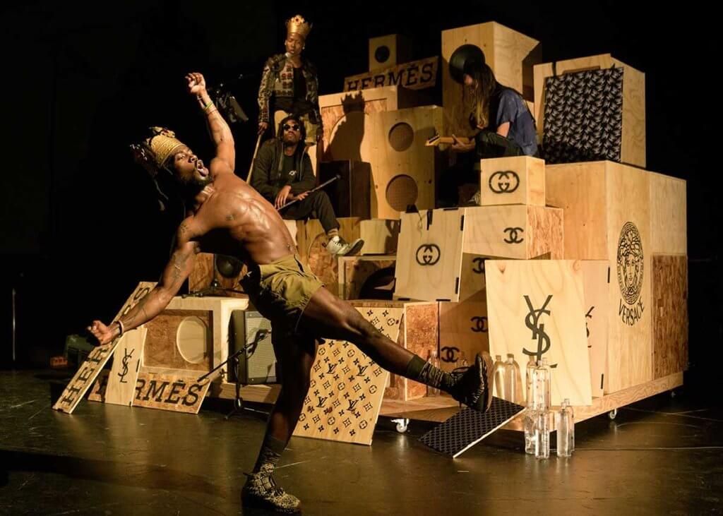 A dark skinned athletic man balancing on one leg in a cross lateral, while two dark skinned people and a light skinned person are mounted on a sculpture of a makeshift sound system created by Kara Walker.