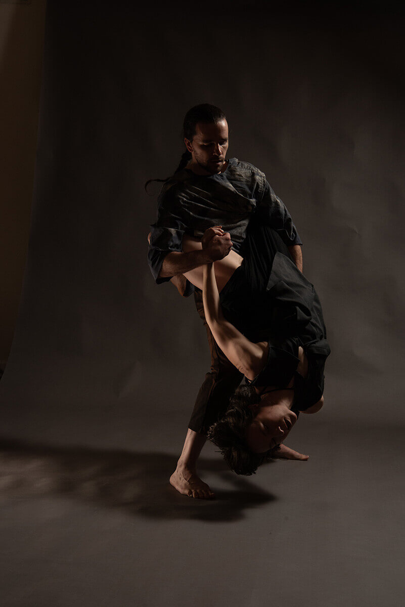 Two white folks (anna thompson and taylor knight) are partnering in a dimly lit room with a grey background. Anna hangs upside down, their dark hair is wildly suspended in time.They have their legs wrapped around taylor’s neck, and are holding their hand to provide support. Taylor has their hair slicked back into a ponytail braid, they are looking down towards anna with their left hand providing support on their back.