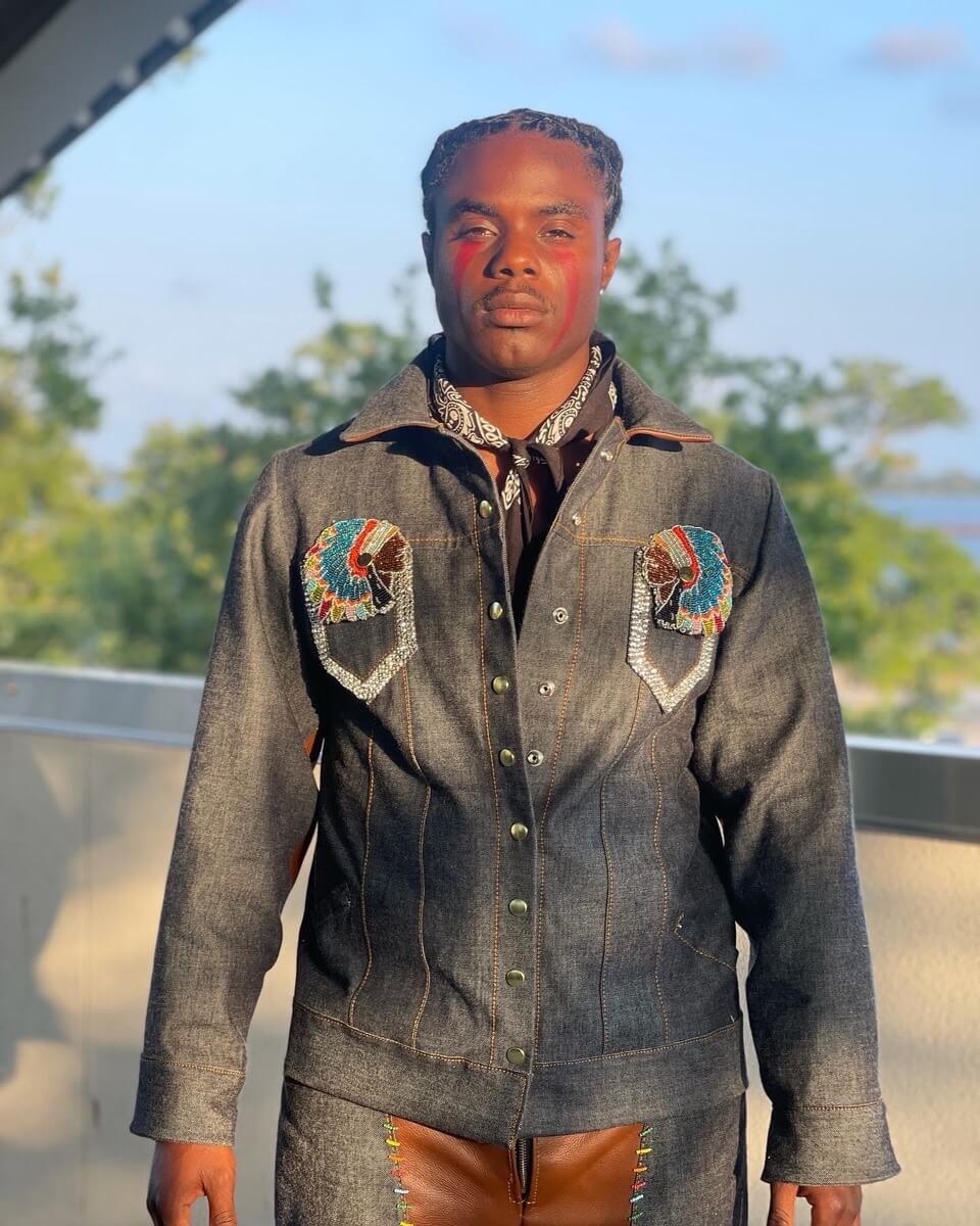 A Masking Indian poses for the camera on a balcony in a decorated denim jacket and trousers. He has a black bandana around his neck and braided hair that is pulled away from his face. Two rhinestoned patches depicting the heads of darkskinned chiefs in colorful war bonnets are situated symmetrically on his chest.