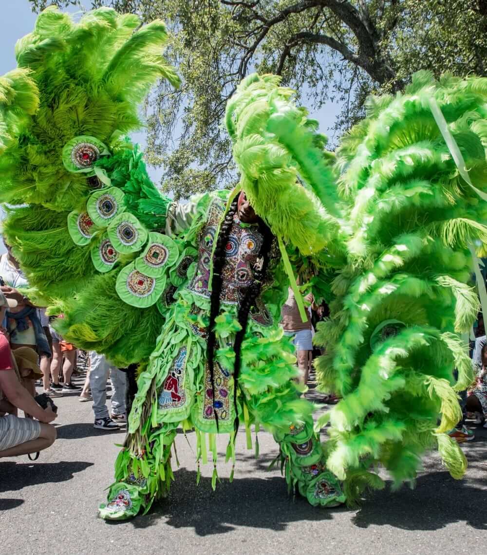 A Masking Indian dances on a sunny, crowded street with his arms wide-open in a green feathered multicolor rhinestoned suit. His wing is spread on the left side of the photo under his feathered arm and his large, feathered war spear is on the right side of the photo in his hand. He wears a round feathered sun hat on his head with a multi-green feathered war skirt around his waist. His head is tilted down and left, covering his eyes with his hat, and his two braids follow the curve of his head.
