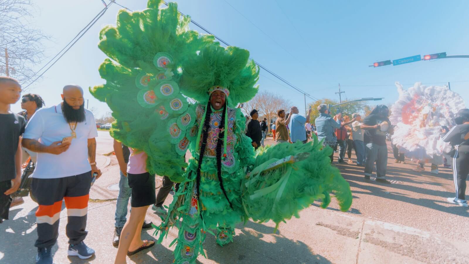 A Masking Indian dances in a green-feathered multicolor rhinestoned suit. His wing is spread on the left side of the photo under his feathered and raised arm, and his war spear points forwards on the right side of the photo in his hand. He wears a round feathered sun hat on his head with a multi-green feathered war skirt around his waist. His eyes are fixed on the camera and his mouth is open mid-yell.