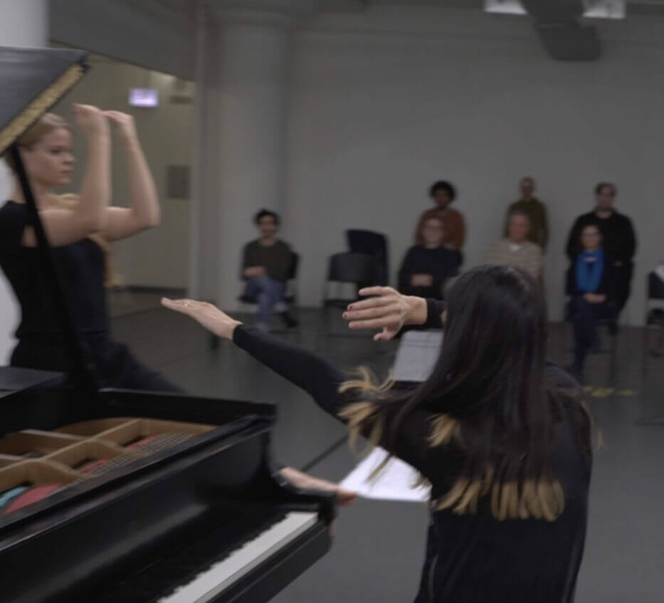 The view is from behind a woman sitting at a grand piano. Her left forearm is in front of her eyes, palm facing out, as her right hand is fully extended. A woman with a blonde ponytail faces right. Their arms are bent at the elbow in front of their head, palms facing in and hands in a curved shape. A blurry audience is watching them.