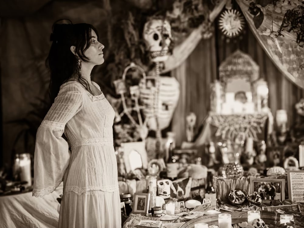 Sydney Calderon A photo is a side profile of a light-skinned woman standing before an elaborate Dia de Muertos altar lit with candles. She is a Chicana woman who wears a long-sleeved white dress; her hair is pulled back with loose curls surrounding her face. The image is a black-and-white print.