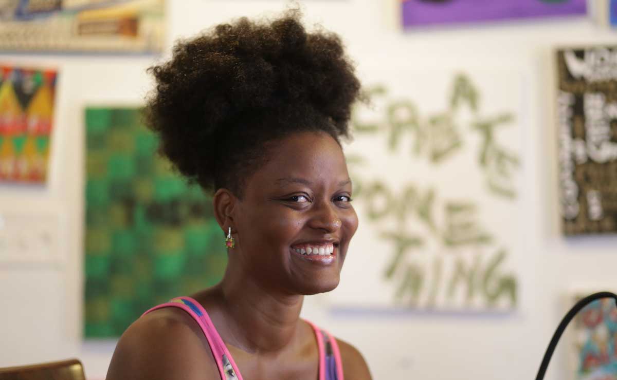 A young Black woman with natural hair pulled up over her head smiles at the camera. Behind her, blurred, is a white wall covered with prints, paintings, and other art.