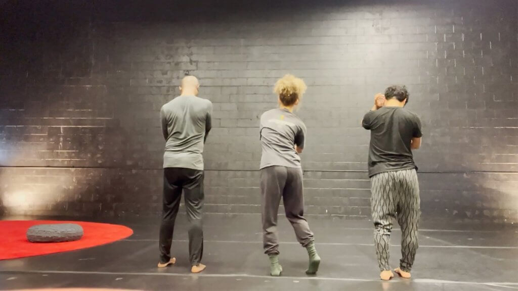 Image shows David Thomson, Anna Martine Whitehead, and Takahiro Yamamoto, all wearing shades of gray with their backs toward the camera. They are all barefoot, Anna has on green socks. They are in a stark gray/black room. There is a red round carpet with a small round cushion on the left side of the performers.