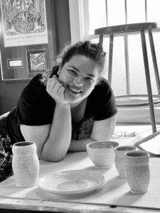 Black-and-white photo of a light-skinned woman in a black T-shirt, head resting in her hand as she looks down over a set of dinnerware.