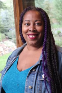 A light-brown skinned woman with twists and some purple twists mixed in. She is in a turquoise shirt and a denim jacket.