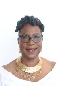 A headshot of a dark-skinned woman with an upswept hairstyle. She is wearing eyeglasses, a white off-the-shoulder top, brass Adinkra-symbol earrings, and a brass necklace that has Adinkra symbols hanging from it.