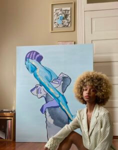 A brown skinned woman seated next to a large-scale painting. One leg is bent upwards with her arm resting on her knee. The painting depicts a female figure facing away from the frame with the back of her bun showing. The colors in the painting are blue, purple, gold, and black.