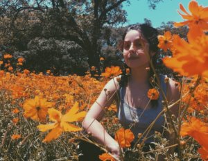 A light-skinned Latinx person with long dark hair in braids is sitting in a field of orange cosmo flowers. They are gazing at the camera with a calm expression. They are wearing a simple gray tank top, a gold chain, large gold hoop earrings, and pink-tinted round sunglasses. There are trees and a blue cloudless sky in the background.