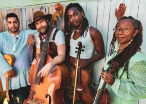 Four musicians are standing in front of a white painted fence. The man to the far left is light-skinned, wearing a long white chemise, and carrying a Renaissance guitar. The man next to him is white and holding an upright bass. He has a brown beard, brown hat, and brown overalls. The person next to him has ebony skin. They are wearing a crop-top t-shirt and holding a viola. Next to them is a woman with tortoiseshell glasses and locs. She is a dark-brown skinned woman in a green, long-sleeved dress, holding a violin.