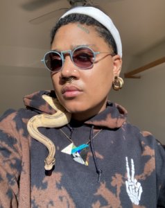 Artist Lorna Williams standing in the sunlight with their pet snake, Crux the Route, sitting on her shoulder. She is wearing a white hat and a black and brown hoodie. Lorna is wearing a handcrafted necklace that she produced.