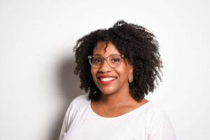 Headshot of Monica, from the shoulders up, wearing a white shirt and red matte lipstick standing in front of a white background.