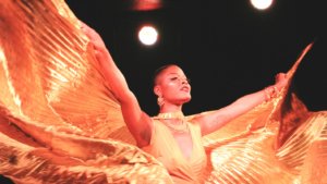 A brown skinned woman mid-movement dancing. Her face is smiling, arms outstretched and holding golden Isis wings, which she is moving back and forth like waves. She is also wearing a golden yellow dress.