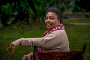 This image is a picture of Tommye Myrick, a milk-chocolate-shade woman, sitting in her director’s chair. She has her back to the camera and looks over her left shoulder toward the camera. The back of the chair says “Director.”