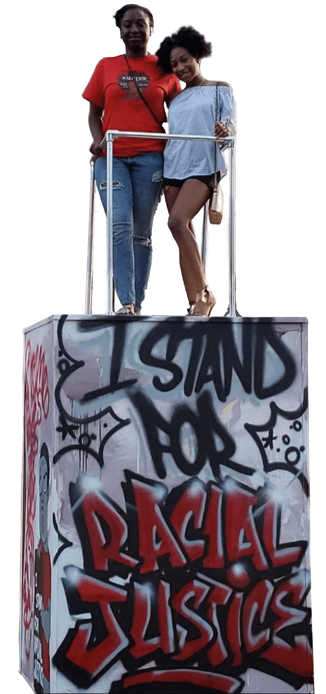 A four feet by four feet wooden pedestal stands approximately six feet high. It's painted in broad, messy brush strokes of grayish-purple and light gray, with graffiti-style lettering painted on top that reads, "I STAND FOR RACIAL JUSTICE," with "I STAND FOR" in black capital letters and "RACIAL JUSTICE" in graffiti tag-style red lettering. The words fill the entire front face of the platform. One side of the platform is partially visible on the left, on which is painted a human figure in red and gray alongside red lettering and white lettering, but due to the angle of the image, this text is unreadable. On top of the pedestal is a viewing area designated by silver metal safety poles. Two Black women stand inside the the viewing area, smiling at the camera as they pose. The woman on the left is wearing a red t-shirt and blue jeans, and the woman on the right is wearing a light blue off-the-shoulder blouse and shorts.