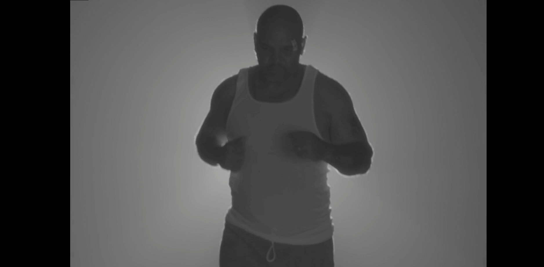 In this still from the documentary "after angola," a man in a white muscle-tee runs toward the camera. He is in semi-darkness with a faint light behind him.