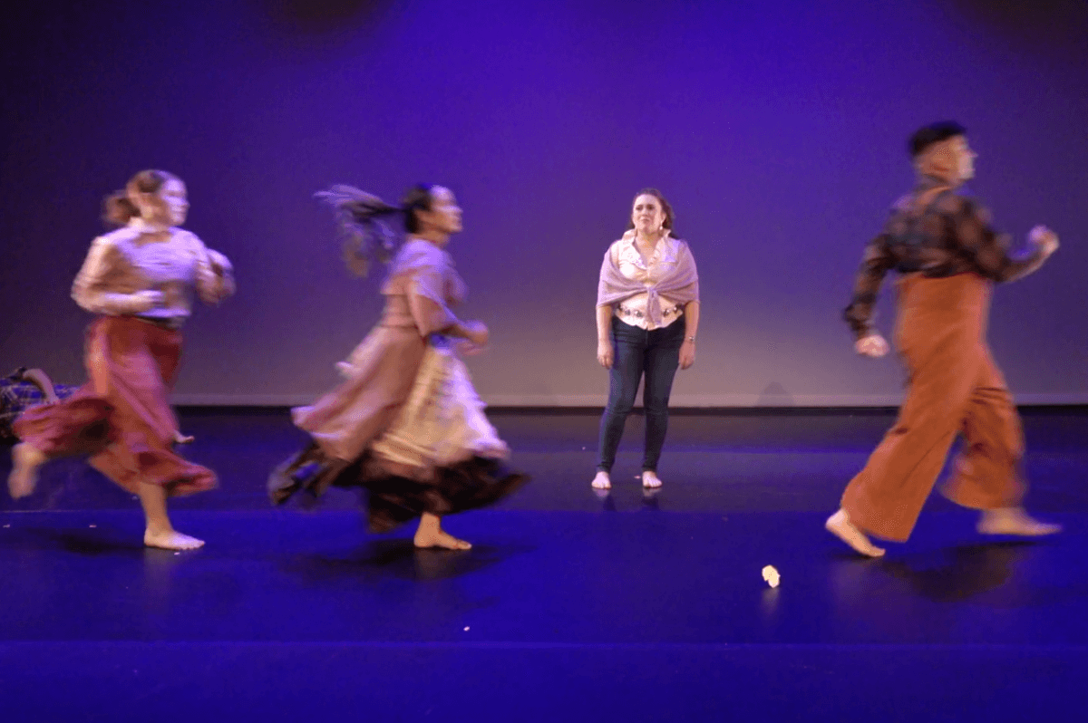 Four dancers perform on a stage lit in blue and purple. Three of the dancers are running across the stage from left to right, and are slightly blurred from the motion. Two of these dancers are women and the other is a man The fourth performer, a woman, stands motionless upstage with her arms by her side, facing the audience.