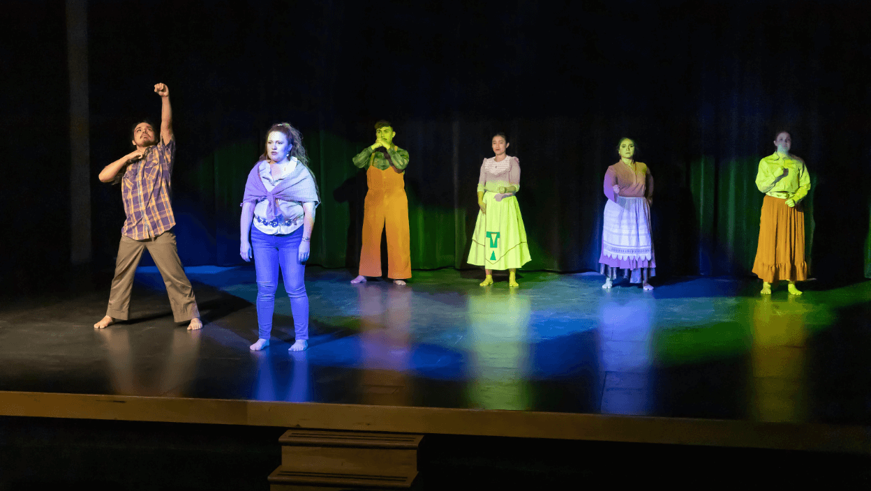Six dancers stand barefoot on a proscenium stage with black curtain backdrop and bright yellow and blue stage lights. In the foreground is a Nuevomexicana woman wearing blue jeans, white button up blouse, purple shawl around her shoulders and a silver concho belt. She looks to the audience and speaks with arms down by her sides. Slightly behind her left shoulder stands a Mexican American man wearing light brown wide leg pants and a purple flannel top with short sleeves. He stares at his left fist as it's raised towards the sky; his right arm is bent with a lifted elbow and hand close to throat. In the background four dancers stand in a horizontal row creating gestures with their arms.