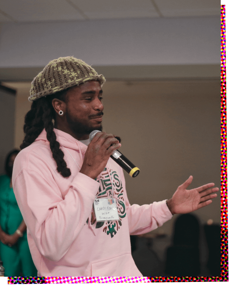 Photograph of Carey Fountain, a Black man dreadlocks pulled back and wearing a greyish-green fuzzy knit cap. He is standing in three-quarter profile facing left, and holds a microphone in his right hand as he gestures with his right hand. He is wearing a light pink cotton hoodie with a decorative green and maroon pattern on the chest.
