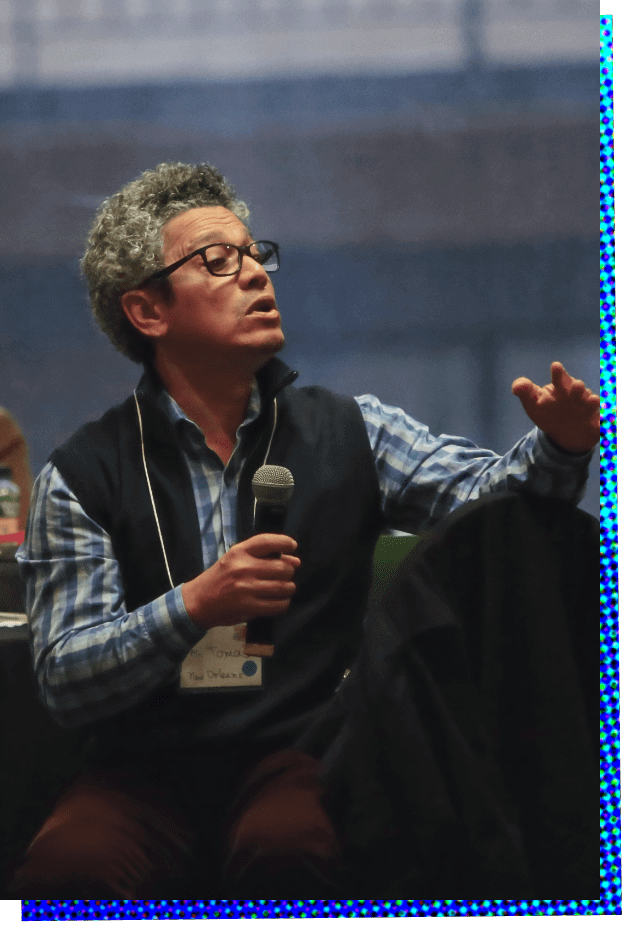 Photograph of Tomas Montoya Gonzalez. A Latino man with closely cropped gray and black hair and wearing glasses leans forward from his chair as he speaks into a microphone that he's holding in his right hand. His left arm is propped on the back of a chair and he's gesturing with this left hand as he speaks. He is wearing a light blue plaid, long sleeve shirt, and a dark blue or black fleece collared vest.