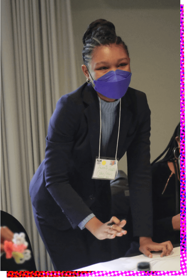 Photograph of Jazzie Jelks, a Black woman. She is slightly bent over a table and holding a pen in her right hand as she looks up and smiles at someone unseen. Her hair is pulled back into a bun, and she is wearing a navy blue pantsuit and blazer, with a light blue blouse underneath. She is wearing a royal blue face mask that appears almost purple in contrast to her suit. 