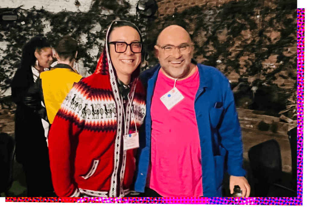 Two men, Daniel Prusarnukul and Antonio Garza, smile for the camera with their shoulders pressed together, suggesting that they have their arms around each other as they pose. On the left, Daniel is wearing a red knit sweater hoodie with a large white band running across the chest that contains a repeating pattern of stylized llamas and shrubs in black. He has black hair that is mostly obscured by the hoodie. He is wearing square framed black glasses with lightly-tinted red lenses, and he has his right hand in the pocket of his pants. On the right, Antonio is wearing a loosely fitting coral pink shirt, over which he has an unbuttoned, deep blue, long sleeve button-down shirt. He is bald and has a closely trimmed light brown and white beard, and wears glasses.