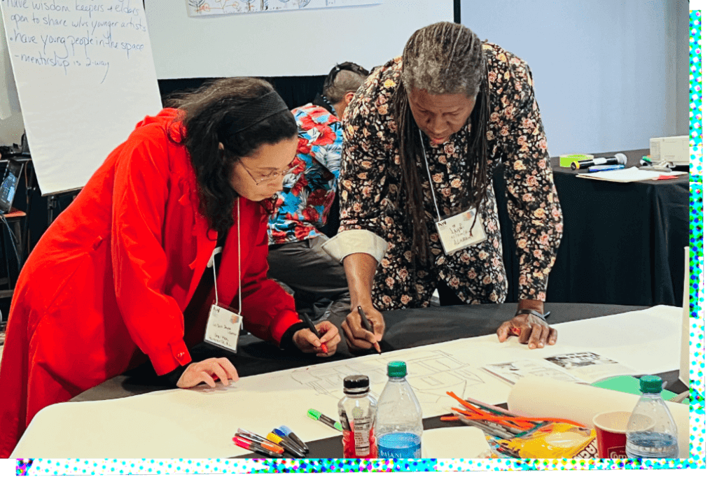LeiSera Jacobs-Leamon and Jahni Moore lean over a table as they work together on a drawing on butcher paper. On the left, LeiSera is wearing a long red overcoat with long sleeves, with a black garment underneath. She has shoulder-length straight black hair held back with a black headband, and wears glasses. Her right hand is pressed on the table to support her, and she holds a red marker in her left hand as she looks at the drawing. On the right, Jahni holds a black pencil in his left hand and is drawing on the paper, and his right hand is pressed on the table to support him. He has long hair, partially gray hair that extends to his lower back, and it is pulled back away from his face and drapes over his shoulders to hang down in front of his shirt. He is wearing a long-sleeved, untucked black shirt patterned with small pink and white roses. To give him more freedom of movement while drawing, the cuff on his left sleeve is folded back to revealing a white inner lining.