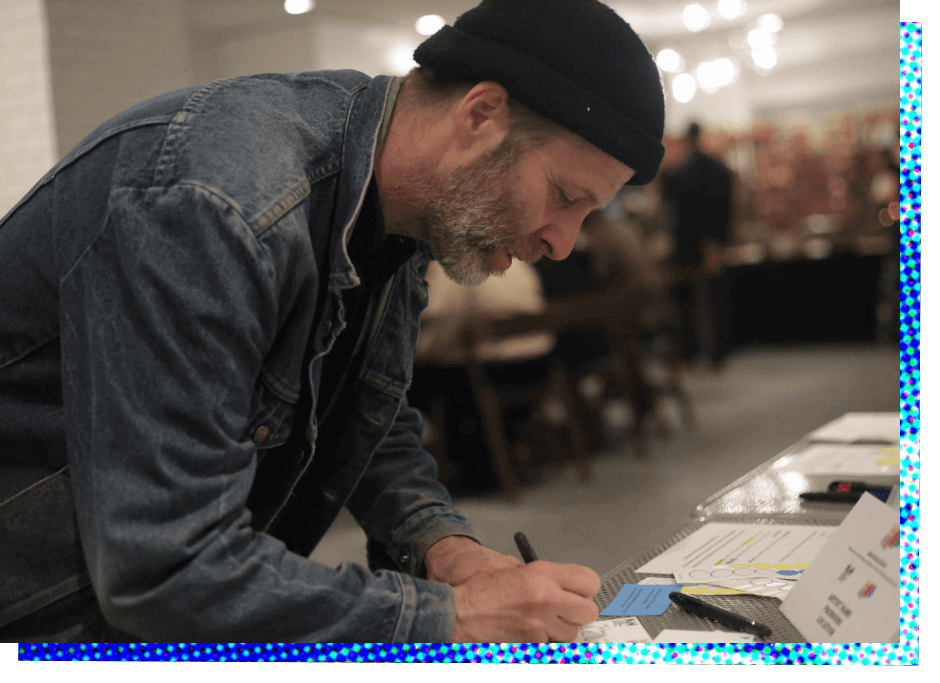 A white man with a trimmed gray and black beard is shown in profile as he leans over a table and fills out a name badge. He is wearing a blue denim jacket and has a tight-fitting black knit cap.
