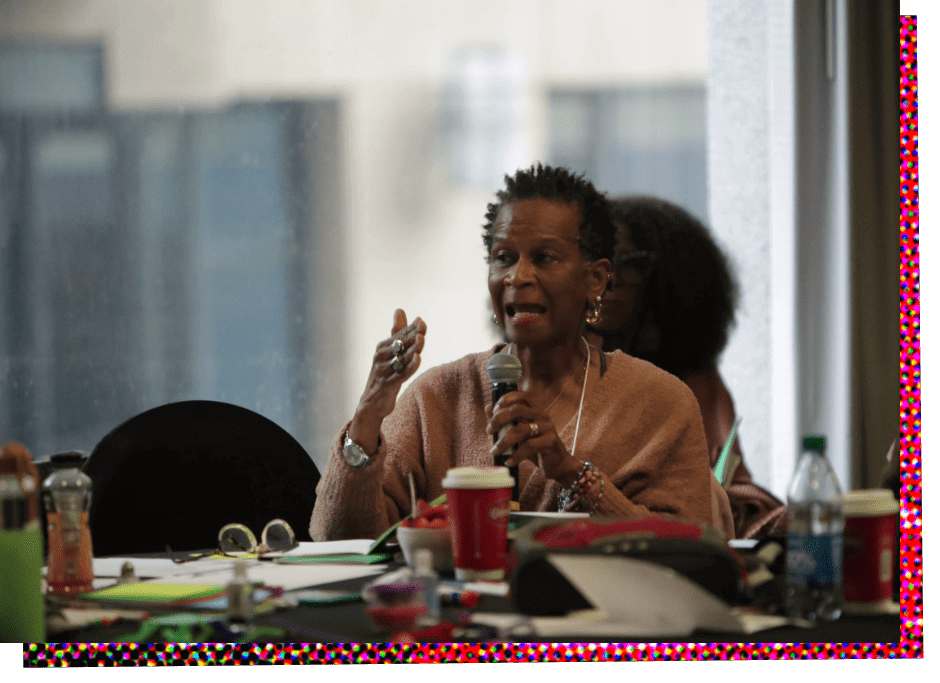 Greer Mendy, a Black woman, sits at a large conference table and speaks into a microphone she is holding in her left hand. She has short black hair and is wearing a light pink or peach, loose fitting blouse with sleeves. The table in front of her is cluttered with papers, writing utensils, cups and bottles.